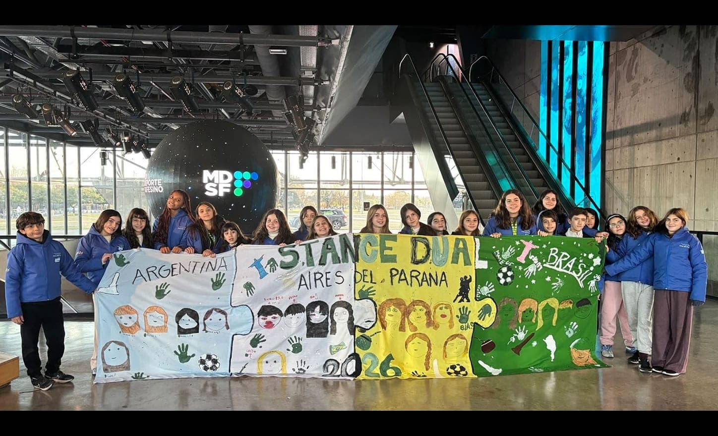 Alunos seguram painel pintado com Argentina, Stance Dual, Brasil e 2026 em um saguão