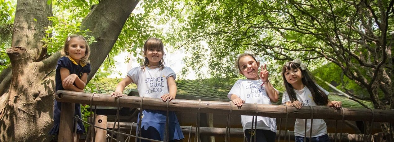 Quatro alunas sorridentes com uniforme da Stance Dual School debruçadas em uma casa na árvore